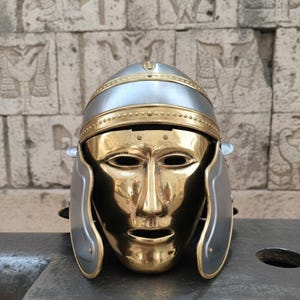 Könnte beinhalten: Ein römischer Helm mit goldfarbener Gesichtsmaske und silberfarbenem Helm. Der Helm hat ein goldenes Band und silberne Wangenklappen. Die Maske hat Augen- und Mundöffnungen.