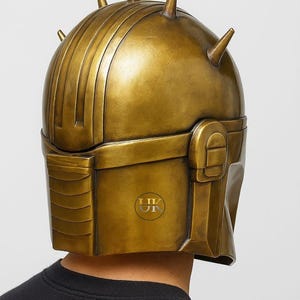 Puede incluir: Un casco dorado con una serie de crestas verticales y varias puntas c&oacute;nicas. El casco tiene una secci&oacute;n rectangular en la parte delantera y una parte superior redondeada. El casco lo lleva una persona que lleva una camiseta negra.