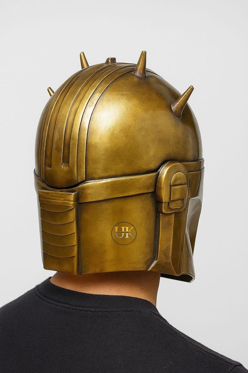 Puede incluir: Un casco dorado con una serie de crestas verticales y varias puntas c&oacute;nicas. El casco tiene una secci&oacute;n rectangular en la parte delantera y una parte superior redondeada. El casco lo lleva una persona que lleva una camiseta negra.
