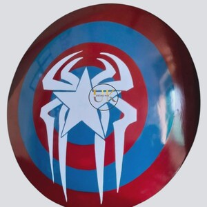 May include: A circular shield in red, white, and blue. The design includes a white star at the centre, encircled by a white spider-like emblem. The shield has a shiny surface.