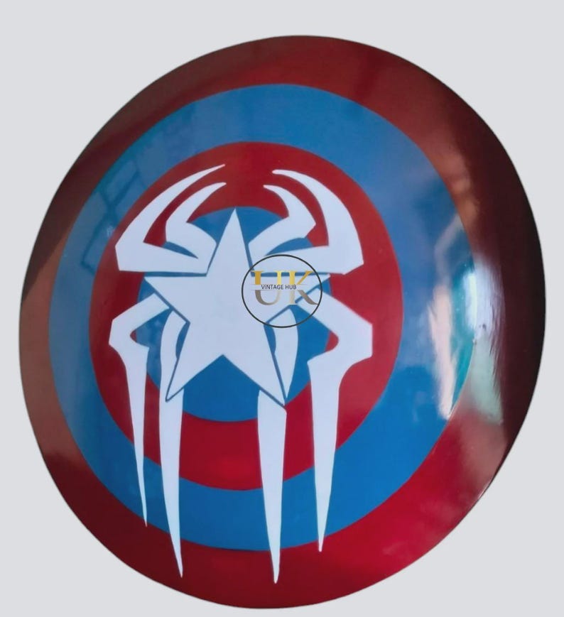 May include: A circular shield in red, white, and blue. The design includes a white star at the centre, encircled by a white spider-like emblem. The shield has a shiny surface.