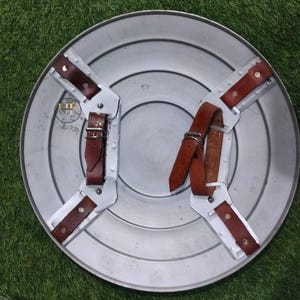 May include: A silver-colored, round shield with a series of concentric circles. The back features brown leather straps and metal supports for gripping. The shield is designed for protection and display.