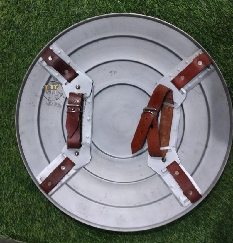 May include: A silver-colored, round shield with a series of concentric circles. The back features brown leather straps and metal supports for gripping. The shield is designed for protection and display.