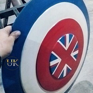 May include: A round shield with a blue outer ring, a white middle ring, and a red center. The center features a Union Jack design. The shield is held by a hand.