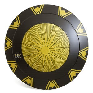 May include: A black and gold shield with a circular design. The centre features a gold starburst pattern, surrounded by a black border with gold geometric shapes. The shield has a glossy finish and a small logo in the lower left corner.