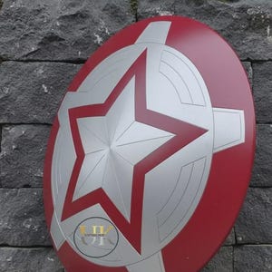Może przedstawiać: Okrągła czerwona tarcza z dużą srebrną gwiazdą pośrodku, otoczona srebrnymi geometrycznymi akcentami i koncentrycznymi liniami. Małe okrągłe logo z napisem VINTAGE HUB jest widoczne w lewej dolnej części tarczy.