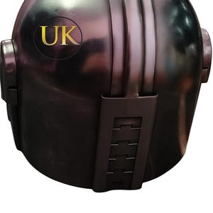 May include: A metallic, dark gray helmet with a rounded top and vertical ridges. The helmet features a gold-colored "UK" emblem and a series of rectangular details on the lower portion. The overall design suggests a futuristic or sci-fi theme.