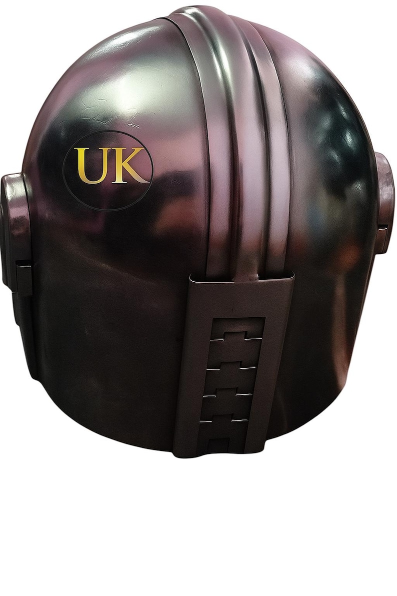 May include: A metallic, dark gray helmet with a rounded top and vertical ridges. The helmet features a gold-colored "UK" emblem and a series of rectangular details on the lower portion. The overall design suggests a futuristic or sci-fi theme.