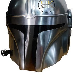 May include: A silver-colored Mandalorian helmet with a sleek, metallic finish. The helmet features a T-shaped visor and a rounded top, with a logo on the upper right side. The design is reminiscent of a space-themed character.