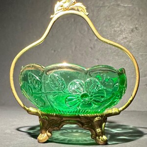 May include: A vibrant green glass bowl featuring a molded floral pattern, held within a gold-toned metal frame. The frame includes a decorative arched handle and three ornate, sculpted feet that support the base of the glass vessel.