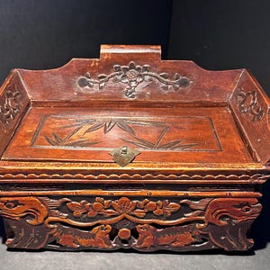 May include: A dark brown wooden box with intricate carvings. The box features a hinged lid with floral and bamboo designs. The base is adorned with carved animal figures and floral patterns. A small metal clasp secures the lid.