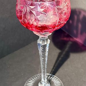 May include: A cranberry red cut-to-clear crystal wine glass featuring intricate starburst and leaf patterns etched into the bowl. The stem is faceted and clear, leading down to a circular base with a radial sunburst design.