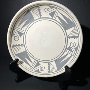 May include: A white ceramic plate with a gray geometric design. The plate features a central circle and a border with triangles, lines, and spiral motifs. The plate is displayed on a black stand.