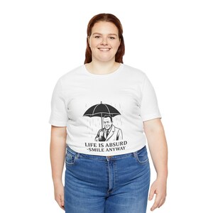 Puede incluir: Camiseta blanca con un dise&ntilde;o en blanco y negro de un hombre con un paraguas bajo la lluvia. El texto "LIFE IS ABSURD - SMILE ANYWAY" est&aacute; impreso debajo. La persona lleva vaqueros azules.
