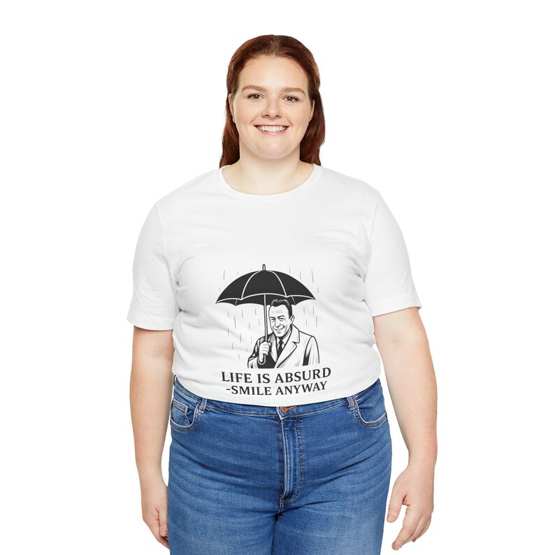 Puede incluir: Camiseta blanca con un dise&ntilde;o en blanco y negro de un hombre con un paraguas bajo la lluvia. El texto "LIFE IS ABSURD - SMILE ANYWAY" est&aacute; impreso debajo. La persona lleva vaqueros azules.