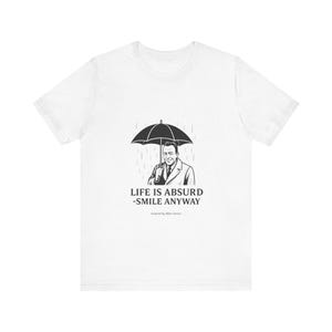 Puede incluir: Camiseta blanca con una ilustraci&oacute;n en blanco y negro de un hombre bajo un paraguas bajo la lluvia. El texto "LIFE IS ABSURD - SMILE ANYWAY" est&aacute; impreso debajo, inspirado en Albert Camus.