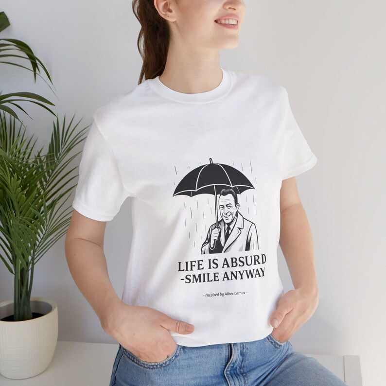 Puede incluir: Camiseta blanca con una ilustraci&oacute;n en blanco y negro de un hombre bajo un paraguas bajo la lluvia. El texto "LIFE IS ABSURD - SMILE ANYWAY" est&aacute; impreso debajo, inspirado en Albert Camus.