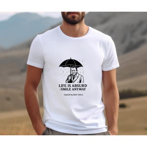 Puede incluir: Camiseta blanca con una ilustraci&oacute;n en blanco y negro de un hombre bajo un paraguas bajo la lluvia. El texto "LIFE IS ABSURD - SMILE ANYWAY" est&aacute; debajo de la imagen, con "Inspired by Albert Camus" debajo.