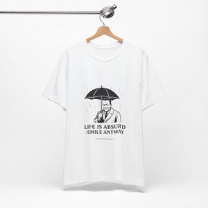 Puede incluir: Camiseta blanca con un gr&aacute;fico en blanco y negro de un hombre bajo un paraguas bajo la lluvia. El texto "LIFE IS ABSURD - SMILE ANYWAY" est&aacute; impreso debajo. La camiseta est&aacute; colgada en una percha de madera.