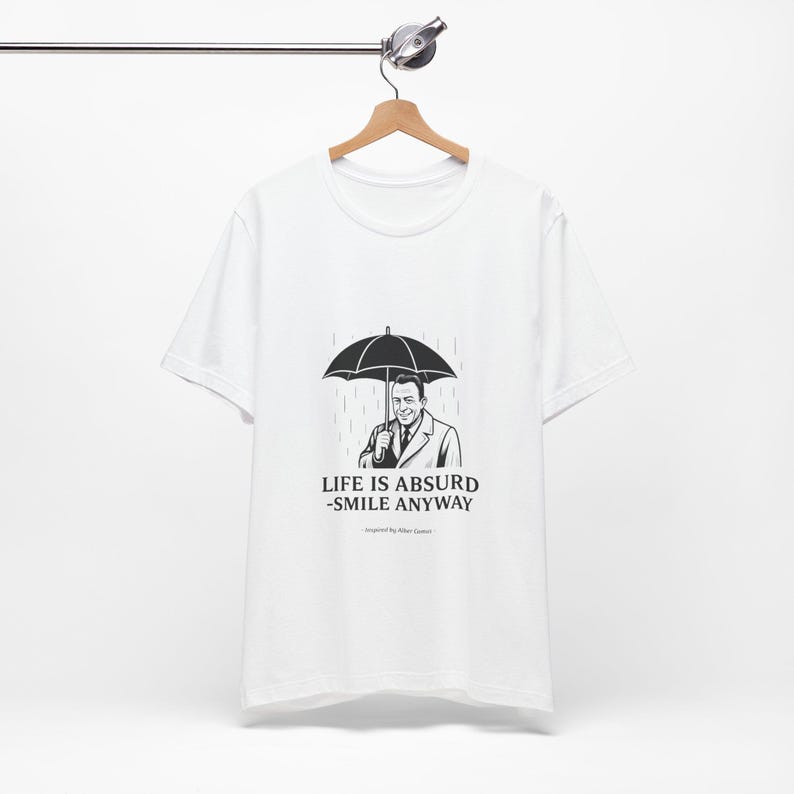 Puede incluir: Camiseta blanca con un gr&aacute;fico en blanco y negro de un hombre bajo un paraguas bajo la lluvia. El texto "LIFE IS ABSURD - SMILE ANYWAY" est&aacute; impreso debajo. La camiseta est&aacute; colgada en una percha de madera.