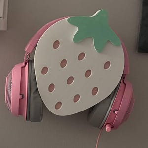 以下が含まれることがあります： 白いイチゴ型の壁掛けに、ピンク色のゲーミングヘッドセットが吊り下げられています。ヘッドセットはグレーのイヤーカップとピンクのヘッドバンドを備えています。イチゴの台座にはピンクの種子のディテールと緑色の茎があります。