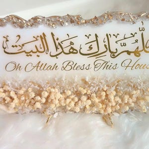 May include: A rectangular white resin plaque with gold metallic edges, featuring Arabic calligraphy and the English text "Oh Allah Bless This House." The base is decorated with a horizontal arrangement of small, dried cream-colored flowers.