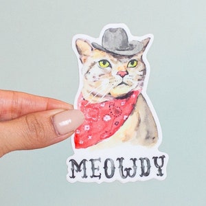 May include: A watercolor illustration of a cat wearing a cowboy hat and bandana. The cat is looking directly at the viewer with a serious expression. The text "MEOWDY" is printed below the cat.