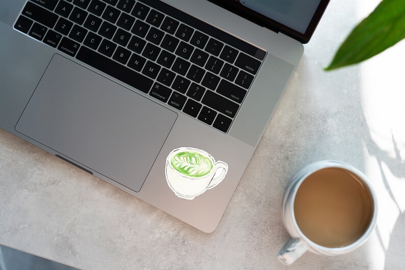 Pu&ograve; includere: Un computer portatile argentato con un adesivo bianco di un latte matcha verde nell'angolo in alto a destra. Una tazza di ceramica bianca con caff&egrave; marrone &egrave; su una superficie bianca.