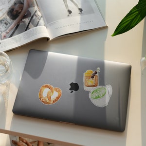 Pu&ograve; includere: Un computer portatile con tre adesivi ad acquerello sul coperchio. Gli adesivi sono di un pretzel, un cocktail e una tazza di caff&egrave;.
