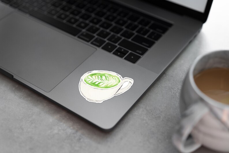 Pu&ograve; includere: Una tazza di ceramica bianca con un design di latte matcha verde su un computer portatile.