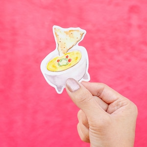 Może przedstawiać: Biała ceramiczna miska wypełniona żółtym guacamole, przykryta jednym chipsami tortilla. Miska ma brązową krawędź i trzy nóżki.