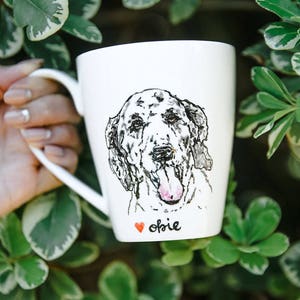 Może przedstawiać: Biały ceramiczny kubek z czarno-białą ilustracją psa. Kubek ma napisane imię "Obie" pod twarzą psa, z małym czerwonym sercem. Kubek jest trzymany przez osobę.