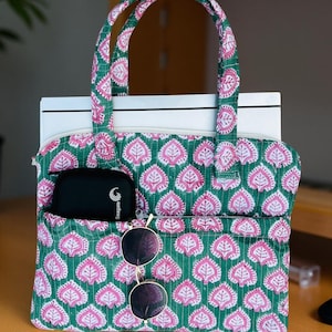 May include: A green and pink patterned tote bag with a leaf design. The bag has a zippered top, two handles, and a front pocket holding a black case and sunglasses. The bag is sitting on a wooden surface.
