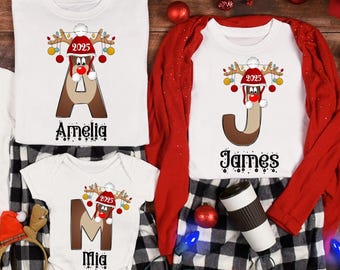 May include: A set of white Christmas family matching shirts featuring reindeer designs, personalised with names and the year 2025. The shirts are paired with black and white plaid pajama bottoms. A red cardigan and festive decorations complete the scene.