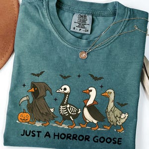 May include: Teal-colored t-shirt with a Halloween-themed graphic. The design features four cartoon geese dressed in spooky costumes, including a grim reaper, skeleton, vampire, and a zombie goose, with the text "JUST A HORROR GOOSE".