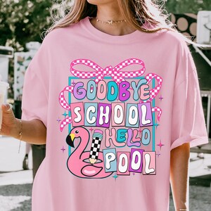 Könnte beinhalten: Ein hellrosa T-Shirt mit einem Grafikdruck mit dem Text GOODBYE SCHOOL HELLO POOL. Das Design enthält einen rosa Flamingo, einen Bleistift mit Karomuster und eine karierte Schleife, alles eingerahmt von einem hellblauen rechteckigen Rand.