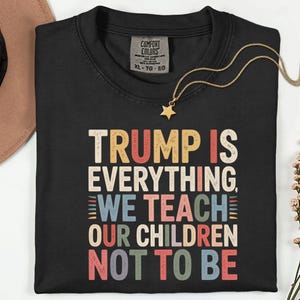 Puede incluir: Camiseta negra con el texto "TRUMP IS EVERYTHING, WE TEACH OUR CHILDREN NOT TO BE" en varios colores. La camiseta tiene una etiqueta Comfort Colors y es talla XL.