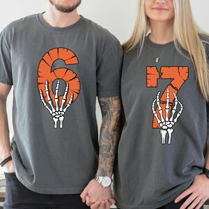 May include: Two gray t-shirts with orange numbers 6 and 7, each with a skeleton hand graphic. The shirts are likely for a couple or group, perfect for Halloween or a themed event. The shirts are made of a soft material.