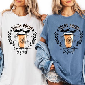 Könnte beinhalten: Weiße und blaue T-Shirts mit einem Halloween-Motiv. Das Design zeigt den Text "Hocus Pocus" über einer Kaffeetasse mit einem Spinnennetz, umgeben von Fledermäusen, Spinnen und einem Lorbeerkranz. Der Text "I need coffee to focus" steht darunter.