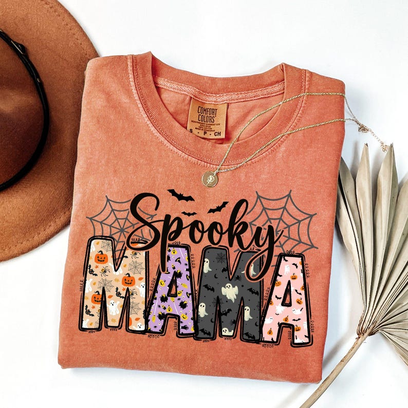 May include: A coral-colored t-shirt with the words "Spooky MAMA" in a Halloween-themed design. The word "Spooky" is in black script above the word "MAMA". The letters of "MAMA" are filled with Halloween patterns. A gold necklace is draped on the shirt.