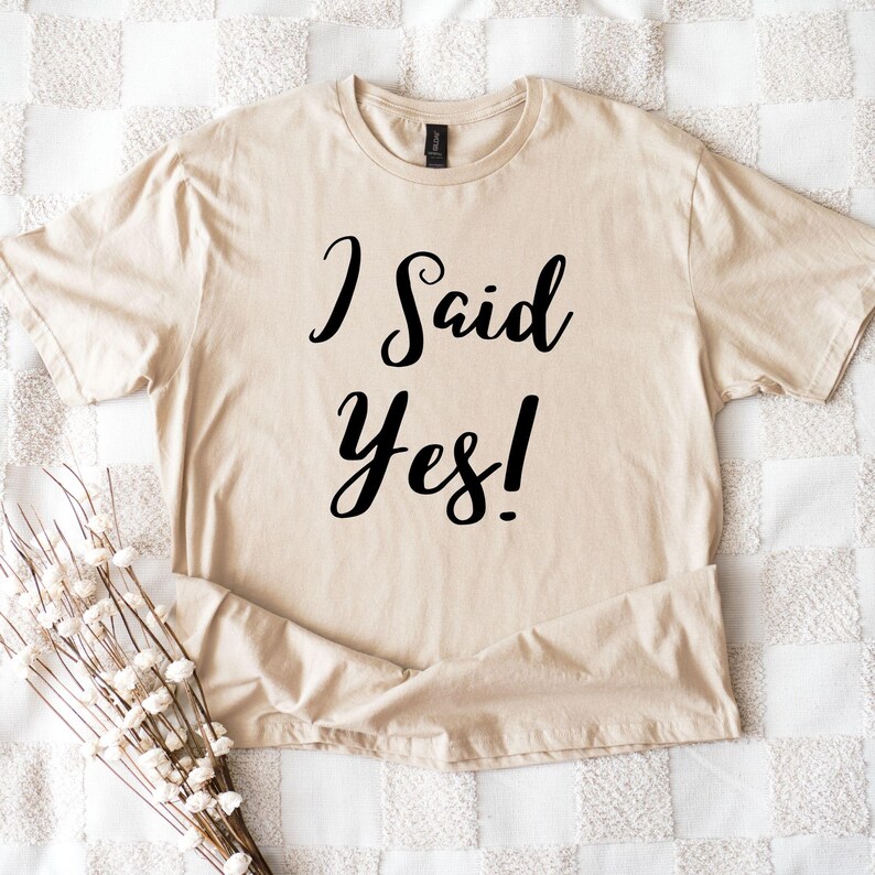 K&ouml;nnte beinhalten: Beiges T-Shirt mit dem Text "I Said Yes!" in schwarzer Schrift. Das Shirt ist gefaltet und auf einer wei&szlig;-beigen karierten Oberfl&auml;che platziert. Ein Strau&szlig; wei&szlig;er Blumen befindet sich in der linken unteren Ecke.
