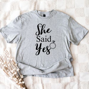 K&ouml;nnte beinhalten: Hellgraues T-Shirt mit dem Text "She Said Yes" in schwarzer Schrift, begleitet von einer Ringgrafik. Das kurz&auml;rmelige Shirt ist aus einem weichen Material gefertigt.