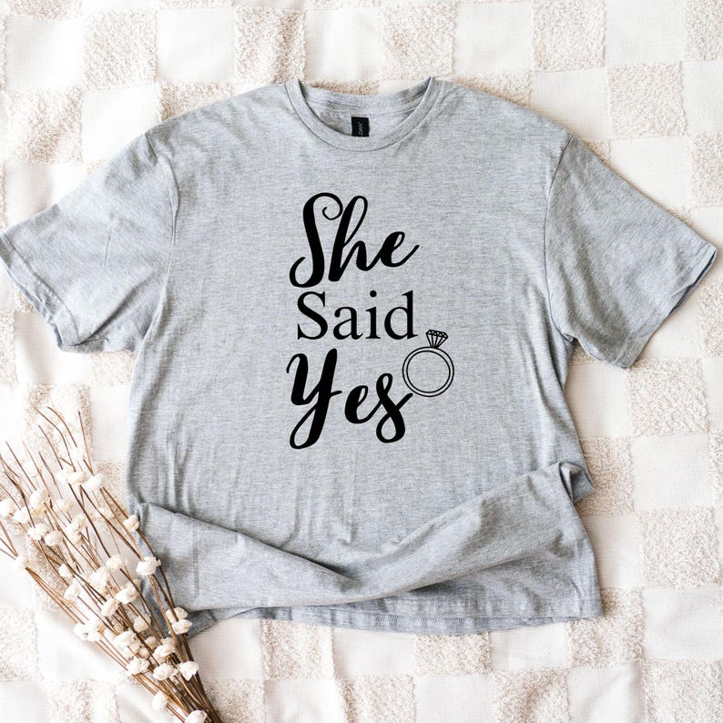 K&ouml;nnte beinhalten: Hellgraues T-Shirt mit dem Text "She Said Yes" in schwarzer Schrift, begleitet von einer Ringgrafik. Das kurz&auml;rmelige Shirt ist aus einem weichen Material gefertigt.