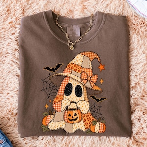 Peut inclure: Un t-shirt marron avec un motif de fantôme portant un chapeau de sorcière en patchwork, tenant un seau citrouille. Le design présente des motifs à carreaux orange et beige, des imprimés floraux, des toiles d'araignée, des chauves-souris et des citrouilles.