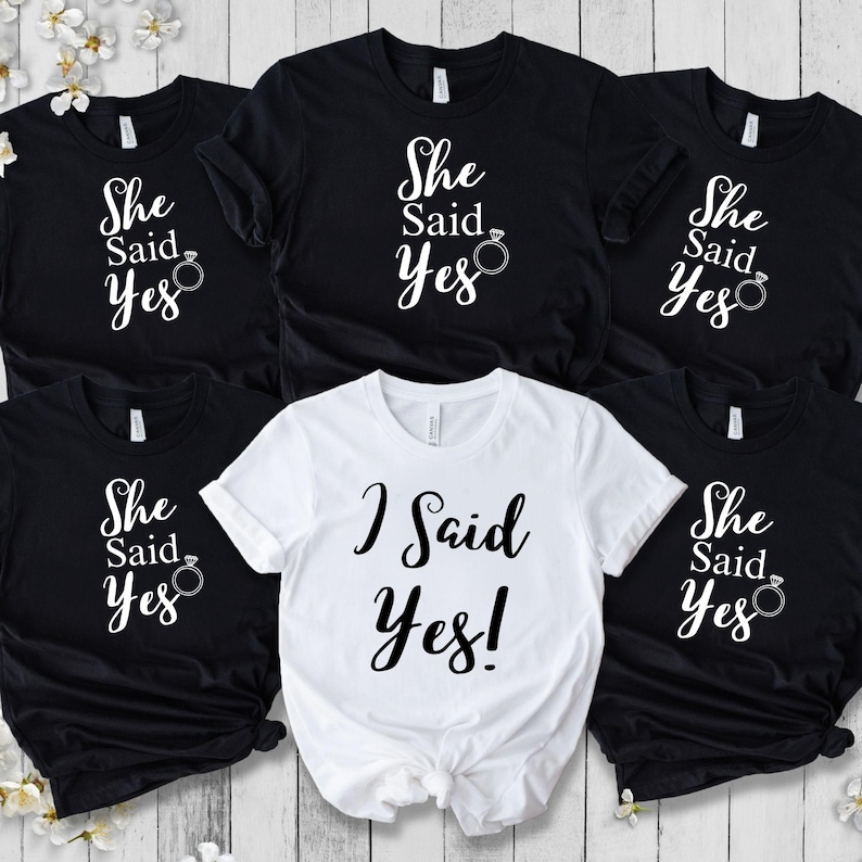 K&ouml;nnte beinhalten: Eine Auswahl an schwarzen und wei&szlig;en T-Shirts. Die schwarzen Shirts zeigen den Text "She Said Yes" mit einer Ringgrafik. Ein wei&szlig;es Shirt zeigt "I Said Yes!" in einer Schriftart. Ideal f&uuml;r einen Junggesellinnenabschied.