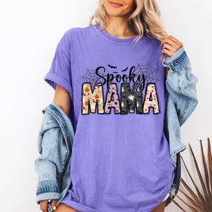 May include: A lilac-colored t-shirt with the words "Spooky Mama" in a Halloween-themed design. The word "Spooky" is in black script above the word "Mama". The letters of "Mama" are decorated with pumpkins, ghosts, and bats.
