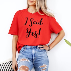 K&ouml;nnte beinhalten: Ein rotes T-Shirt mit dem Text "I Said Yes!" in schwarzer Schrift. Das Shirt wird mit blauen Jeans kombiniert. Das Bild ist bei nat&uuml;rlichem Licht aufgenommen.