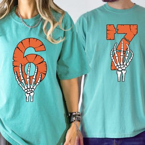 May include: Two teal t-shirts with orange numbers and a skeleton hand graphic. One shirt displays the number 6, and the other displays the number 7. The shirts are made of a soft material and have short sleeves.
