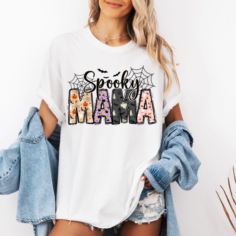 May include: White t-shirt with the text "Spooky Mama" in a Halloween-themed design. The word "Spooky" is in black script above the word "Mama". The letters of "Mama" are filled with Halloween patterns, including pumpkins, ghosts, and bats.