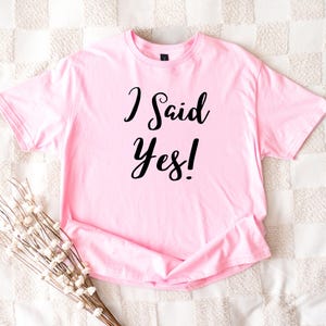 K&ouml;nnte beinhalten: Ein hellrosa T-Shirt mit dem Schriftzug "I Said Yes!" in schwarzer Schrift. Das kurz&auml;rmelige Shirt ist gefaltet, mit einem Zweig wei&szlig;er Blumen in der unteren linken Ecke. Das Shirt liegt auf einer strukturierten, cremefarbenen Oberfl&auml;che.